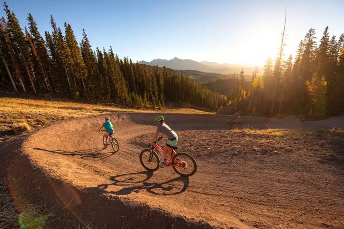 This image captures mountain bikers navigating a curved dirt trail surrounded by tall pine trees, with a majestic mountain range in the background. The dynamic action is set against the wide-open outdoors, suggesting active adventure and family-friendly exploration along scenic trails.