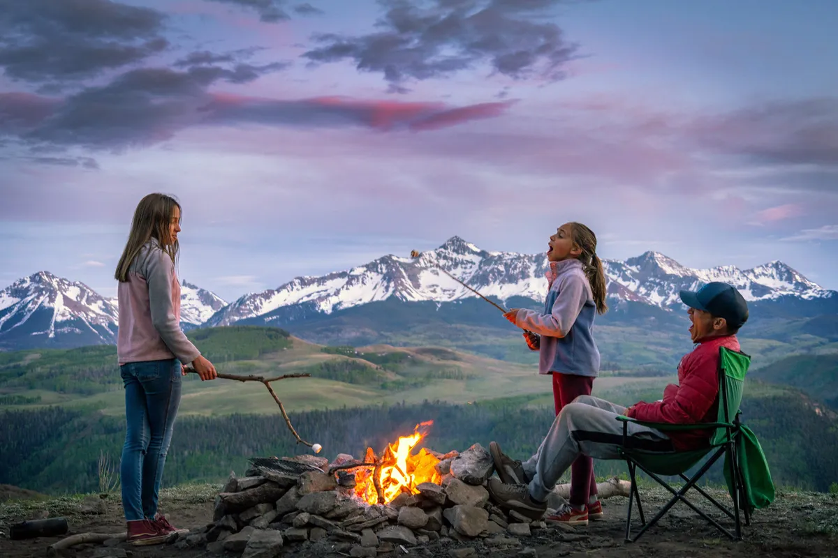 A family scene by a campfire with children roasting marshmallows, framed by a stunning mountain backdrop. The group shares laughter and relaxed moments as dusk falls, highlighting togetherness in a beautiful natural setting.