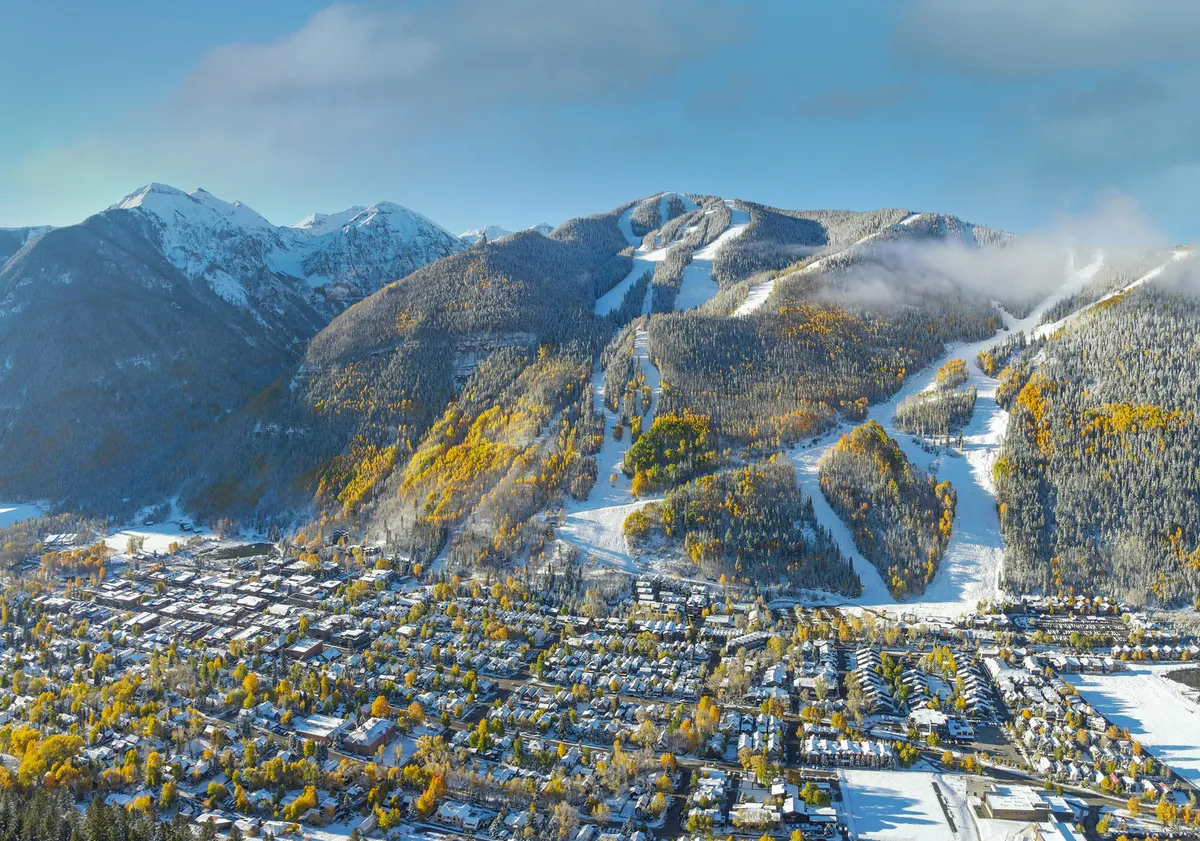 A sweeping view captures snow-capped mountains, winding ski slopes, and the charming town below, all highlighted by splashes of autumn color. The scene blends the excitement of adventure with the tranquil beauty of Telluride's setting.