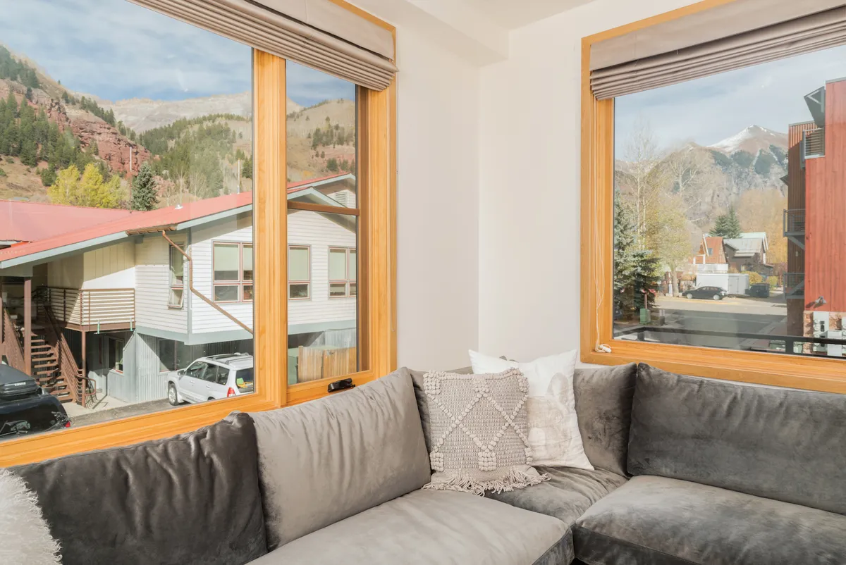 An interior view featuring a cozy sofa with decorative pillows positioned by large windows that frame breathtaking mountain views. The mood is serene and inviting, perfect for quiet moments or informal get-togethers.