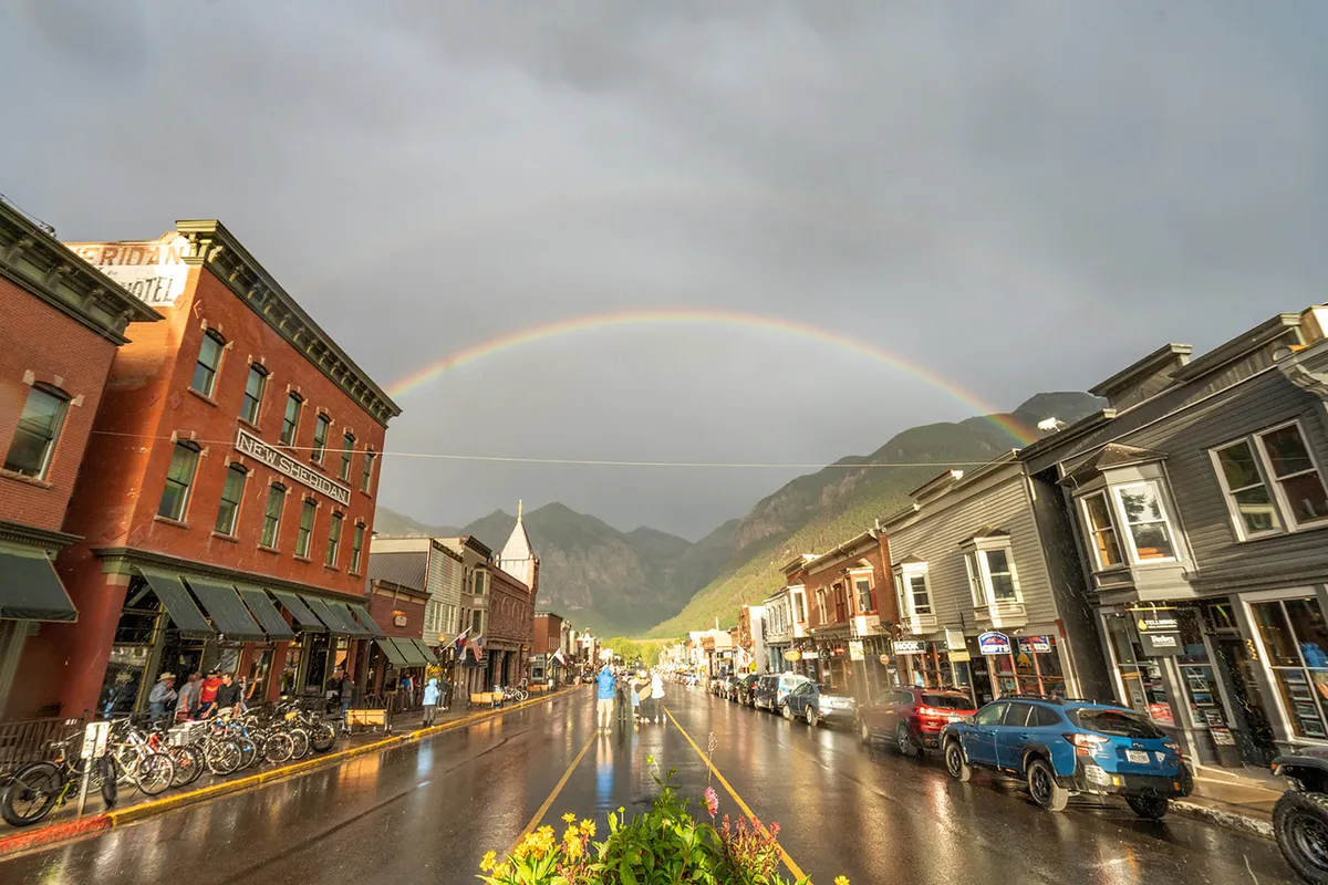 This image captures historic buildings along Telluride's Main Street with a rainbow arching over a wet street. There are bicycles, pedestrians, and hints of outdoor café seating, set against a mountainous backdrop. The lively yet cozy atmosphere showcases the walkability and charm of Telluride's downtown area.