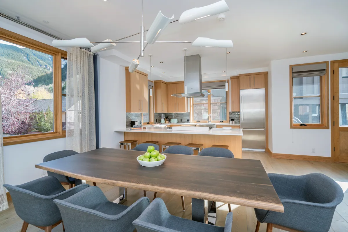 A contemporary, sunlit dining space with a large table, modern kitchen island, and wall-to-wall windows showcasing scenic views. The table is set for a meal and the kitchen is ready for culinary creativity, inviting both intimate and festive gatherings.