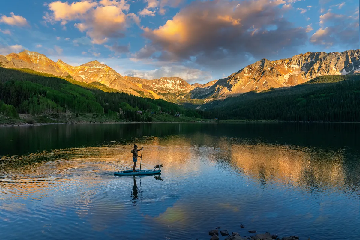 A paddleboarder with a dog enjoys the calm reflections of a pristine lake, framed by mountains and clouds. The tranquil water and expansive scenery suggest peaceful, pet-friendly recreation and the serenity of being immersed in nature.
