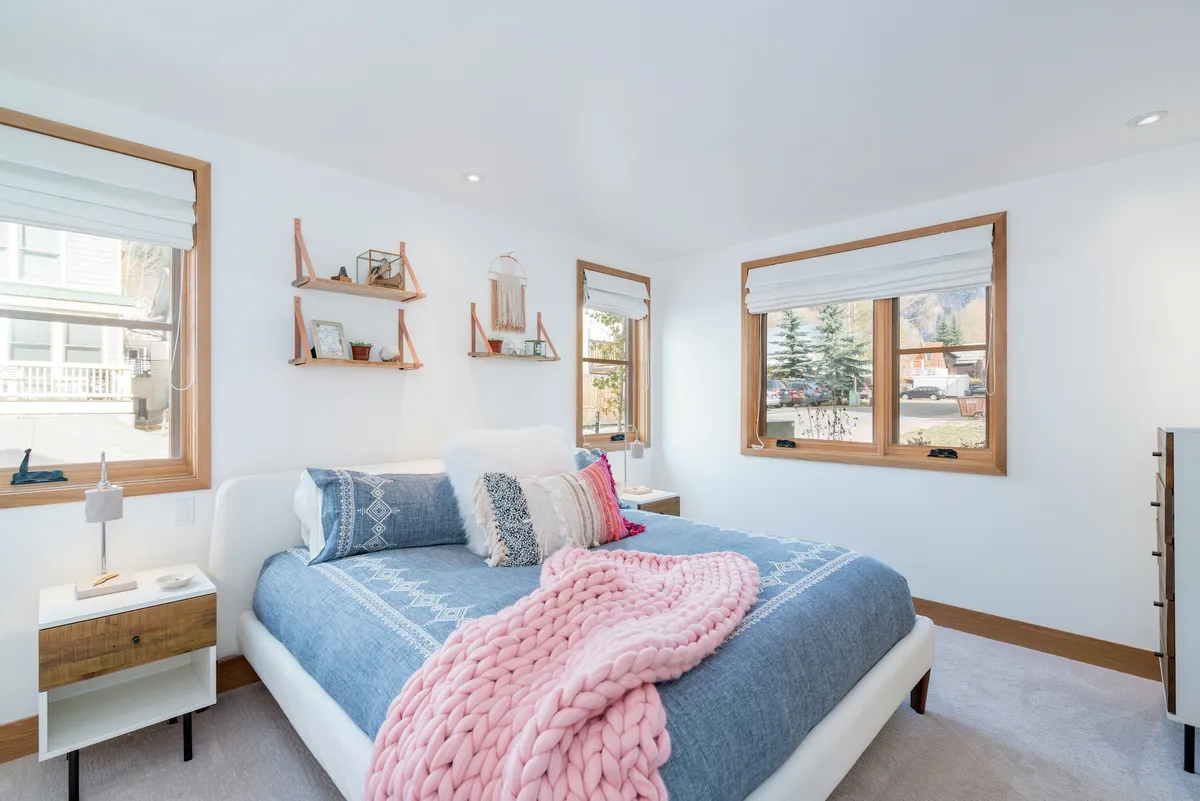 A calm bedroom with a bed, decorative pillows, shelving, and natural light streaming through windows—ideal for a restful personal haven on a lower level.