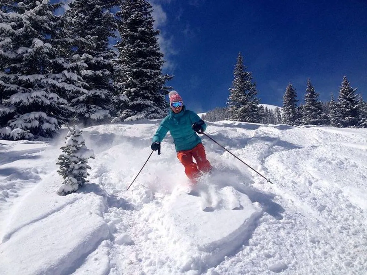 A skier expertly carves down a pristine, snow-covered slope bordered by frosted trees beneath a brilliant blue sky. The scene is vibrant and full of energy, embodying the spirit of winter adventure in Telluride.