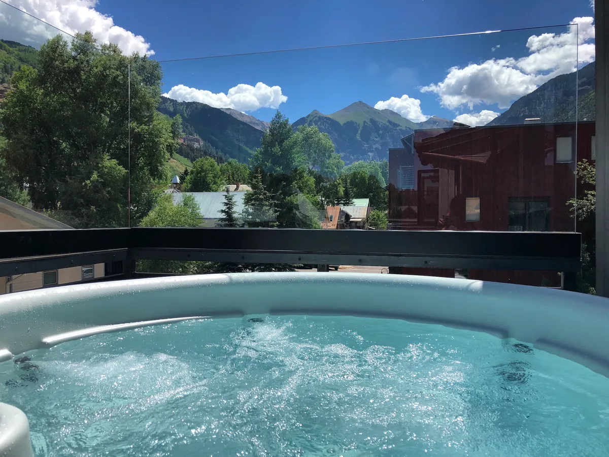 This image features a private hot tub set outdoors with a spectacular mountain view. The scene captures people relaxing in the hot tub, socializing, and soaking in the tranquil atmosphere under a clear blue sky, with sunset and stargazing potential hinted at in the background.