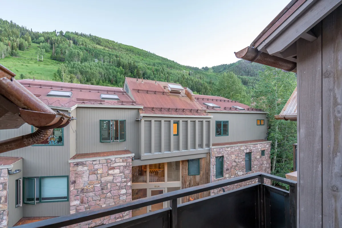 An inviting exterior view of the property, highlighting architectural features, a balcony railing, and the majestic mountain backdrop. The setup suggests a warm, communal space for relaxation and social interaction with nature as a backdrop.