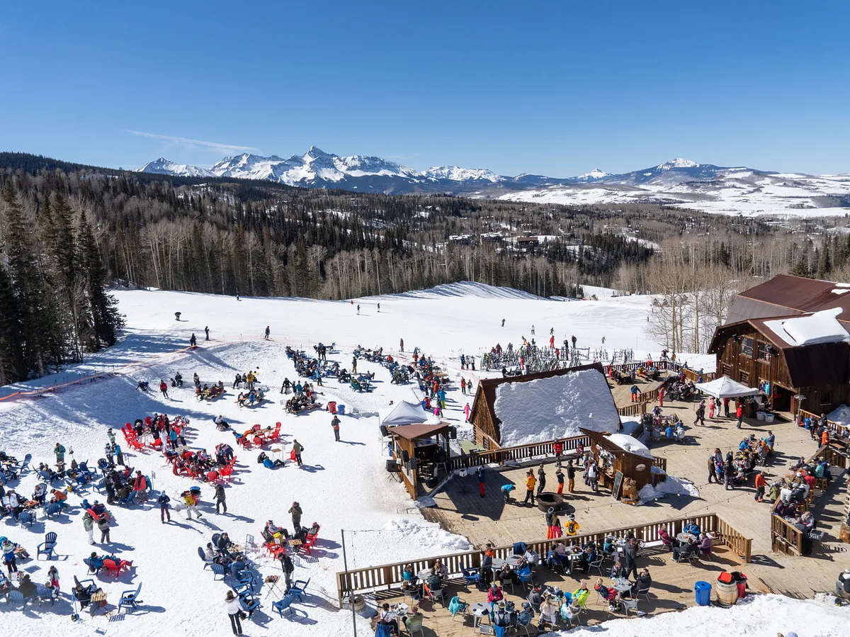 A lively scene featuring a wooden chalet nestled against snow-covered slopes, with people skiing and socializing in an outdoor seating area. The backdrop is a stunning mountain landscape, evoking a sense of adventure and togetherness.